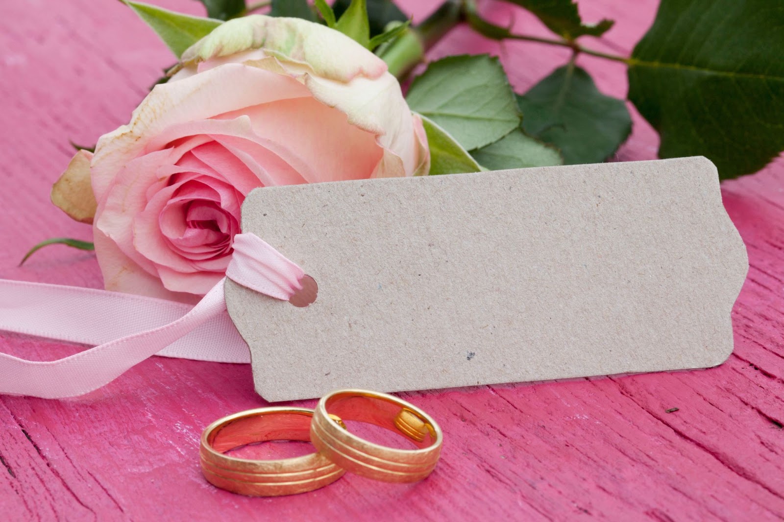 Gold wedding rings next to a pink rose and blank tag on a wooden surface representing personalized touches made possible through wedding competitions.