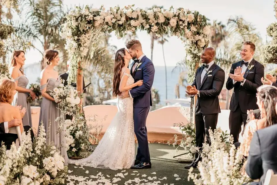 A bride and groom sharing a kiss under a floral arch surrounded by their bridal party — a beautiful moment featured in our affordable wedding guide