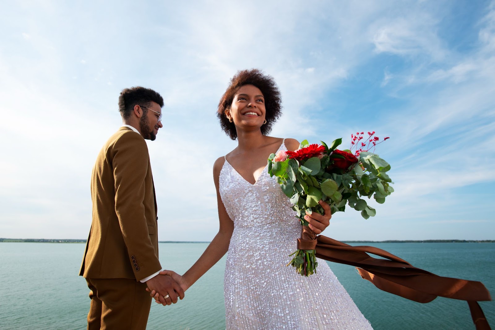 Ethical wedding planning celebration with bride in sequined white dress and groom in mustard suit holding hands by a lake, bride with natural bouquet