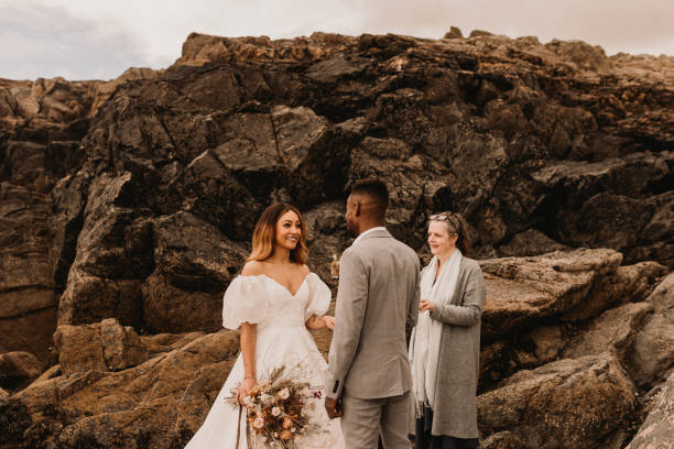A candid shot of a couple and an officiant on a rocky coastline, showcasing a non-traditional venue, a popular choice among current wedding trends.