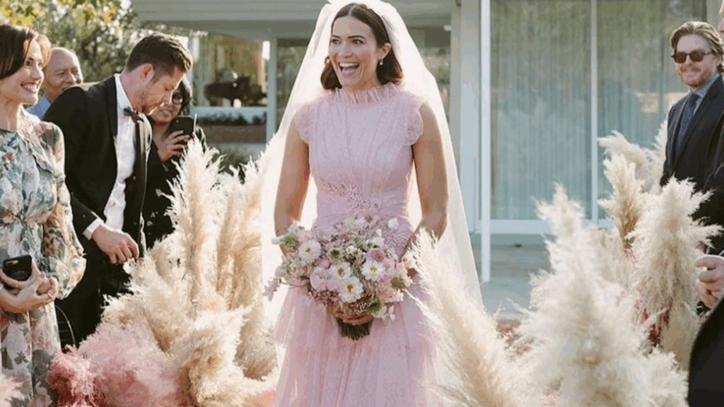 A bride in a unique pink wedding dress laughs joyfully while surrounded by guests, highlighting a vibrant choice in wedding trends.