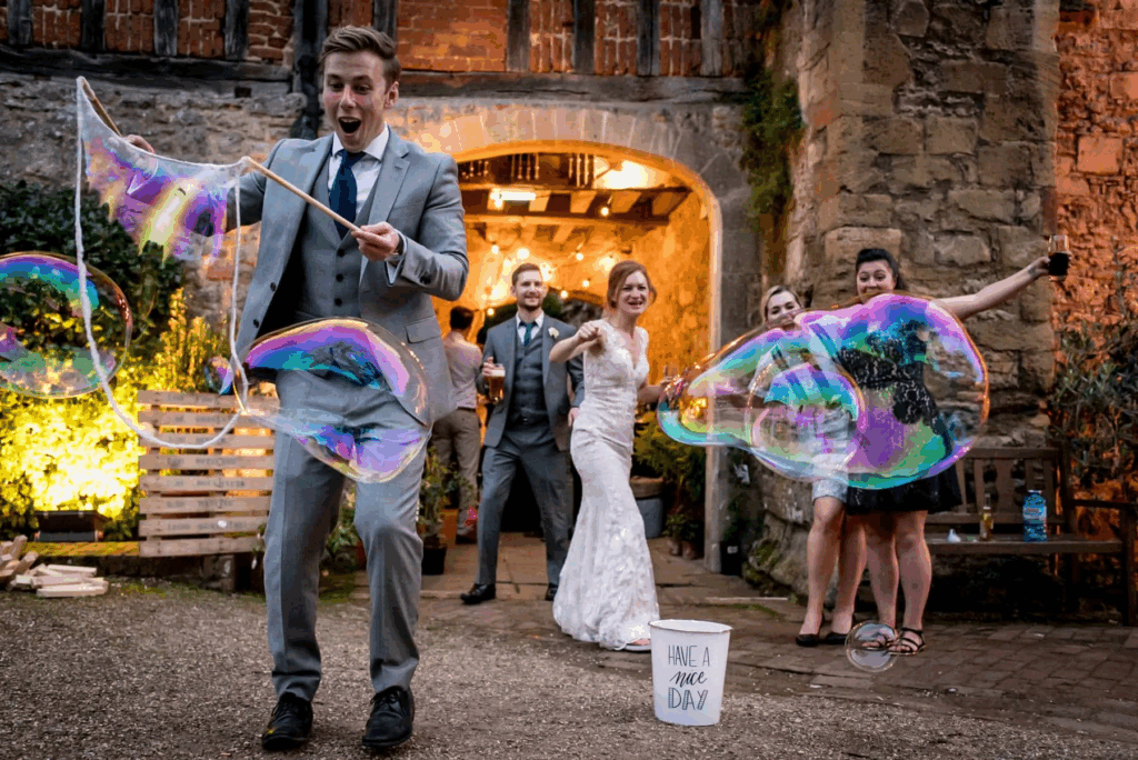 A groom creating large bubbles while guests watch and smile at an outdoor wedding, representing entertaining and interactive wedding trends.
