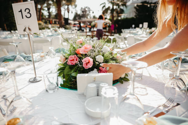 A wedding planner adding a delicate touch to a floral centerpiece on a reception table with a numbered place setting, showcasing attention to detail for the evening's festivities.