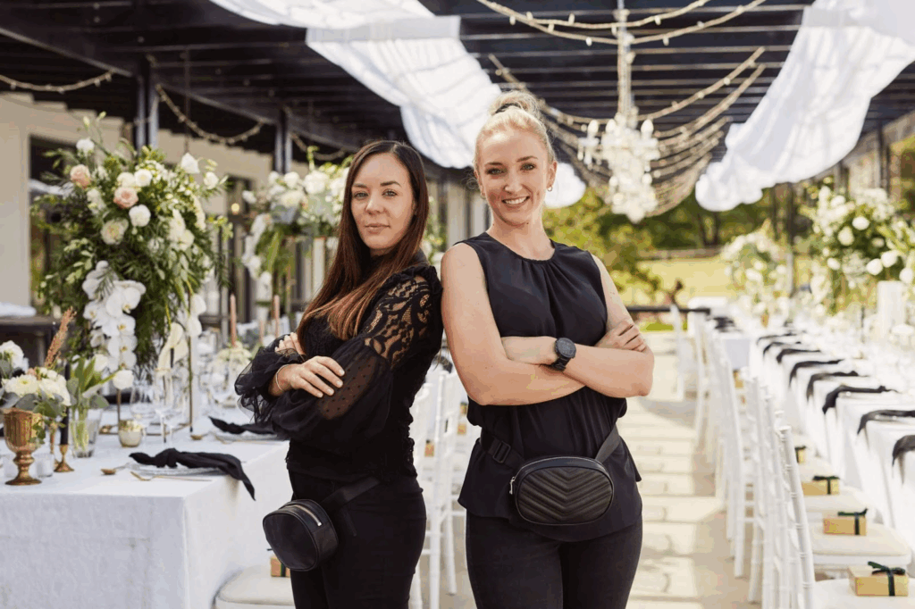  Two wedding professionals stand confidently amidst a beautifully set wedding venue, addressing common myths about the roles of a "Wedding Planner and Wedding Coordinator"